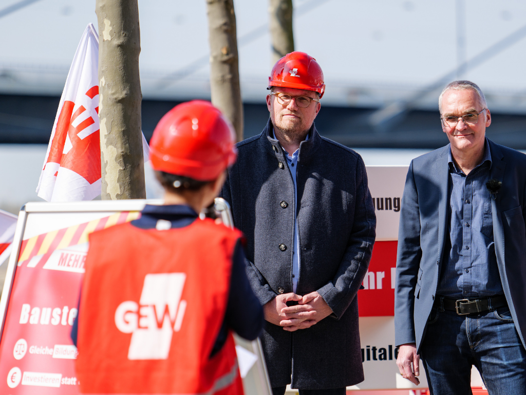Die GEW NRW-Vorsitzende Ayla Çelik hält eine Rede in GEW-Warnweste und mit Bauarbeiterhelm. Jochen Ott und Dietmar Bell (beide SPD) folgen der Rede.