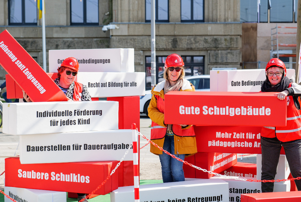 Beschäftigte stehen in der Baustelle Bildung und halten Bausteine mit Forderungen zu guten Schulgebäuden und Entlastung hoch.